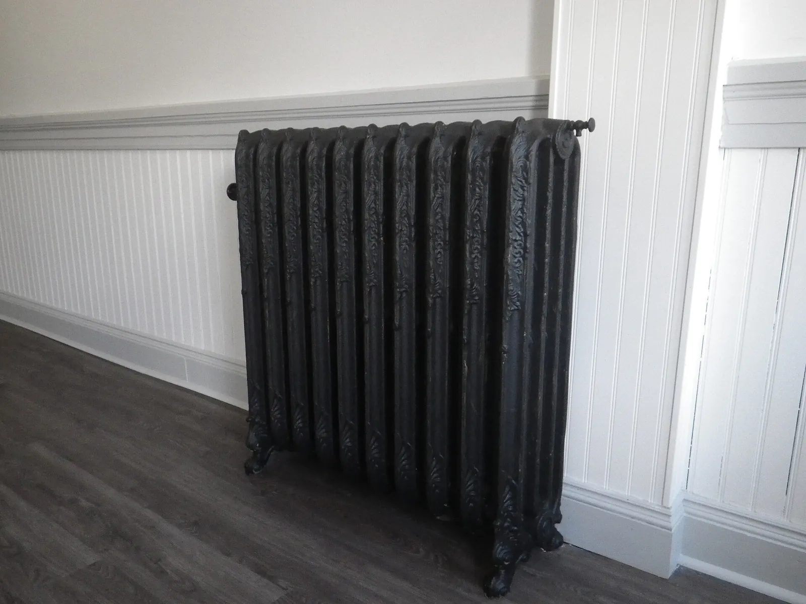Black cast-iron radiator with column sections standing against white wainscoting on a wood floor