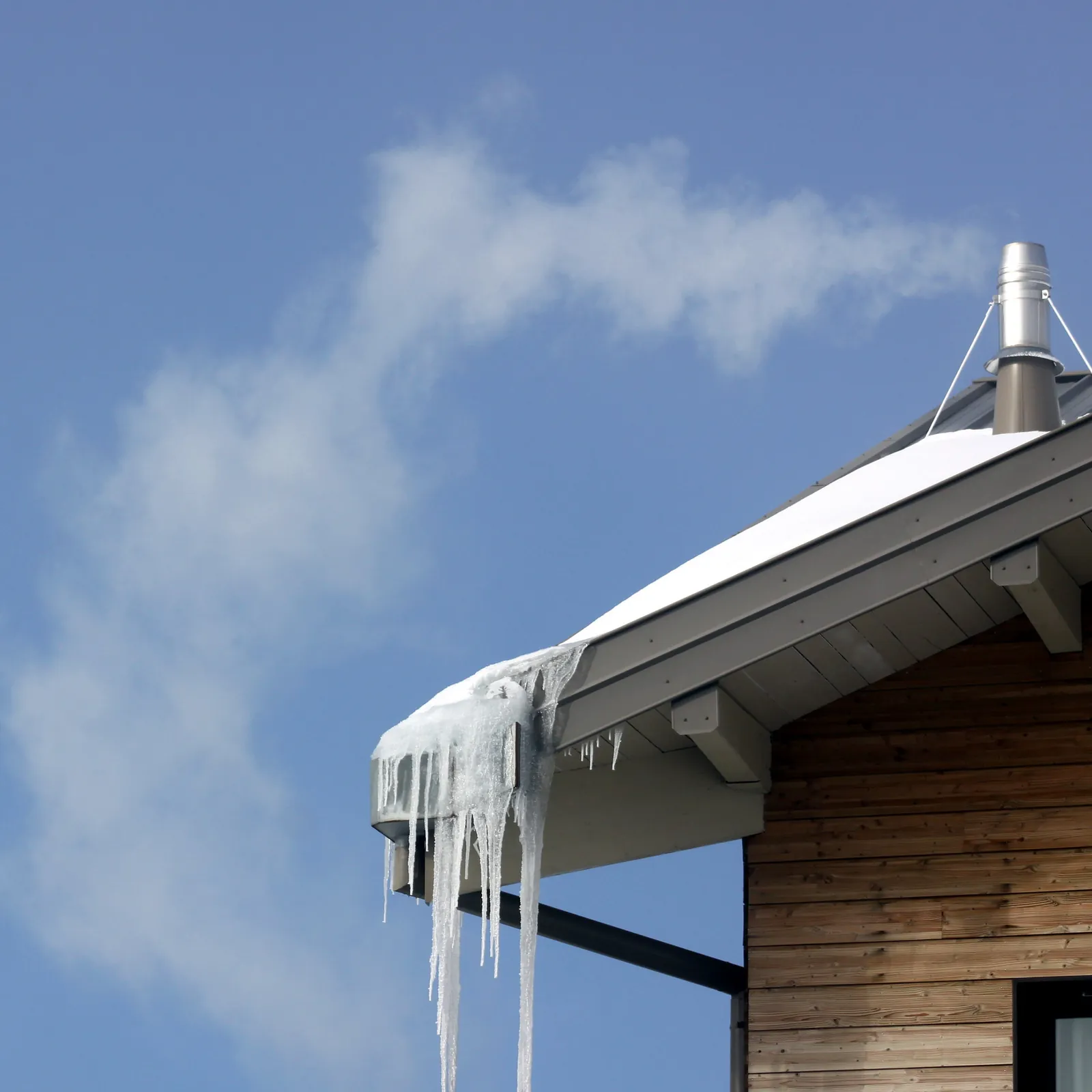 Metal flue pipe venting a vapor plume above a snow-covered roof with long icicles hanging from the gutter