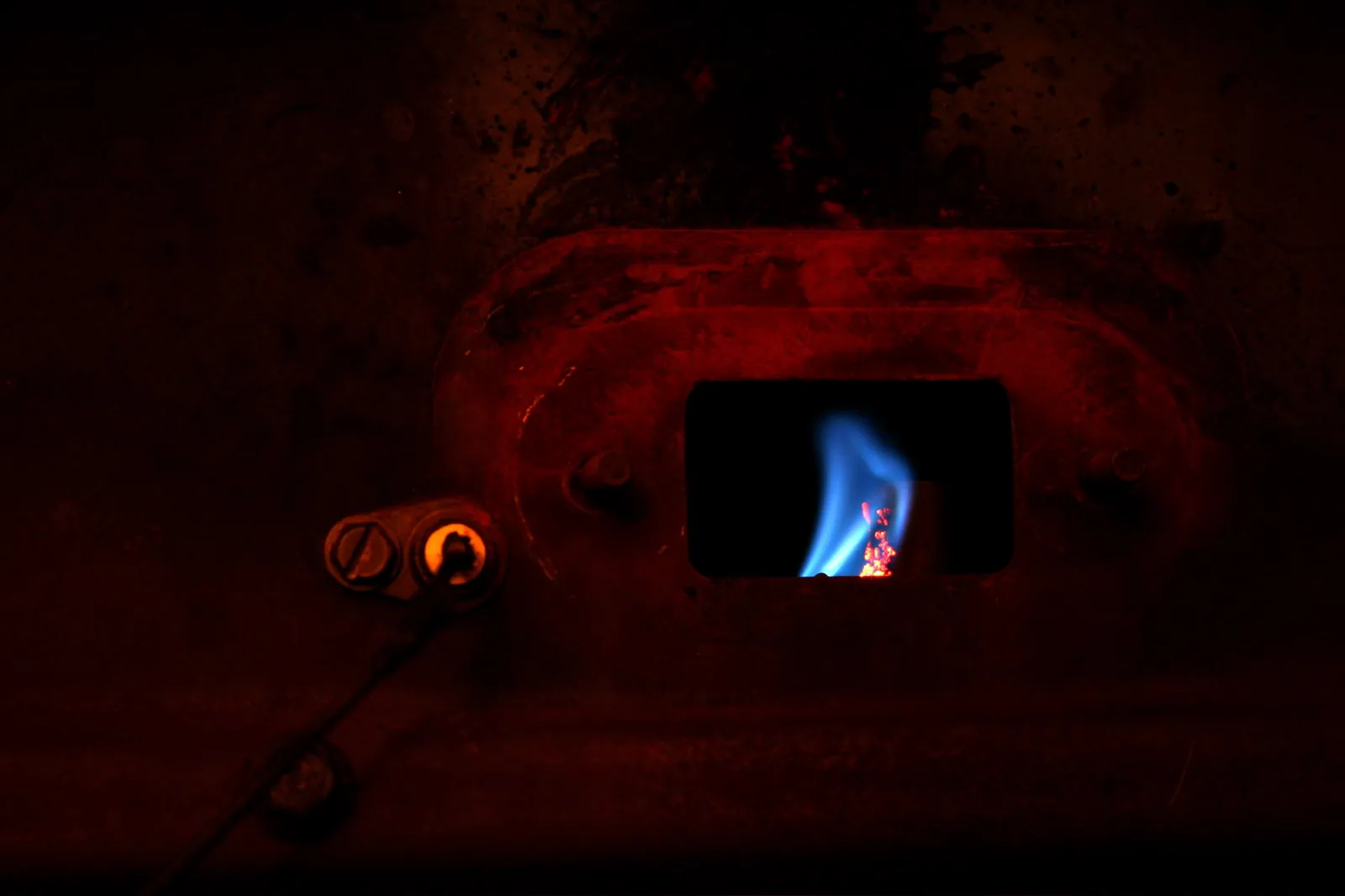 Blue pilot flame seen through the small sight window of a gas heating unit in a dark room