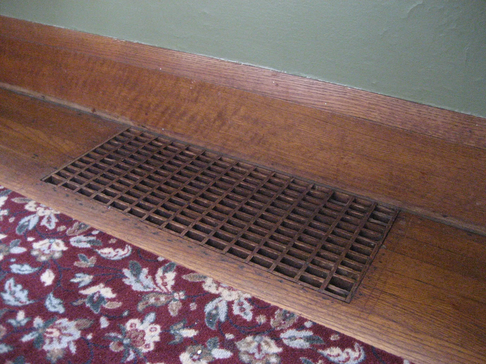 Wooden grid floor register set into an oak floor beside the baseboard, with a red floral rug in front